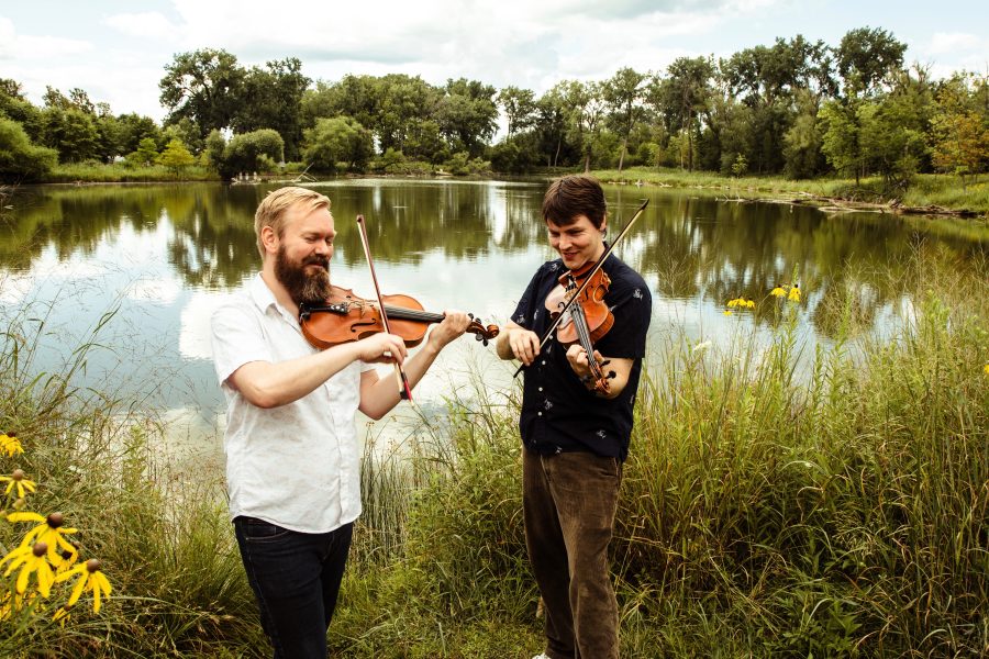 Vidar Skrede, foreground, and Patrick Ahlberg, background, fromt he band Lynx Lynx, stand in fron of a pond with their fiddles.